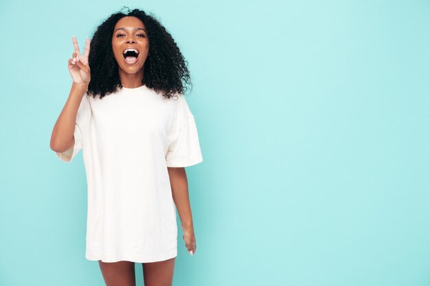 Hermosa mujer negra con peinado de rizos afro Modelo sonriente en ropa de camiseta larga Mujer sexy despreocupada posando junto a la pared azul en el estudio Bronceada y alegre