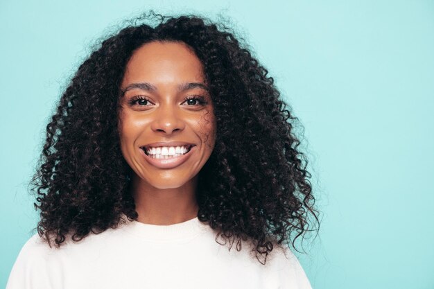 Hermosa mujer negra con peinado de rizos afro Modelo sonriente en ropa de camiseta larga Mujer sexy despreocupada posando junto a la pared azul en el estudio Bronceada y alegre