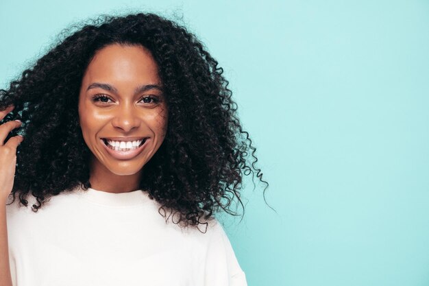 Hermosa mujer negra con peinado de rizos afro Modelo sonriente en ropa de camiseta larga Mujer sexy despreocupada posando junto a la pared azul en el estudio Bronceada y alegre