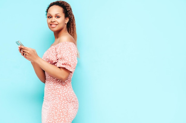 Hermosa mujer negra con peinado de rizos afro Modelo sonriente en ropa de verano de moda Mujer sexy despreocupada posando junto a la pared azul en el estudio Usando aplicaciones de teléfonos inteligentes Mirando la pantalla del teléfono celular