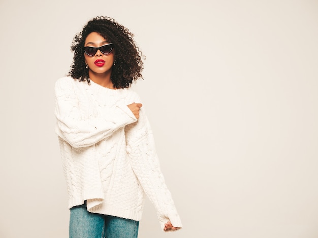 Hermosa mujer negra con peinado de rizos afro. Modelo sonriente en suéter blanco de invierno y ropa de jeans y gafas de sol.