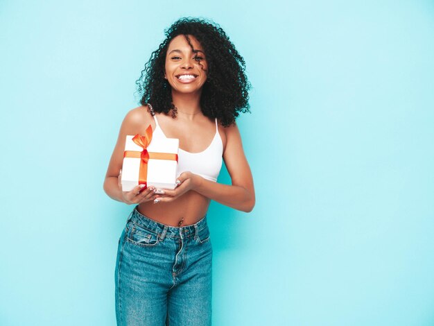 Hermosa mujer negra con peinado de rizos afro Modelo sonriente vestida con ropa blanca de verano Sexy mujer despreocupada posando junto a la pared azul en el estudio Bronceada y alegre Caja de regalo de sujeción aislada