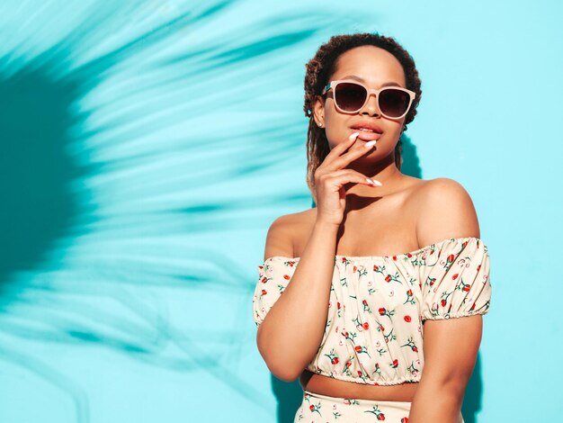 Hermosa mujer negra con peinado de rizos afro Modelo sonriente vestida con ropa hipster de verano Sexy mujer despreocupada posando junto a la pared azul en el estudio Bronceada y alegre En gafas de sol