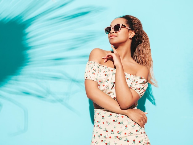Hermosa mujer negra con peinado de rizos afro Modelo sonriente vestida con ropa hipster de verano Sexy mujer despreocupada posando junto a la pared azul en el estudio Bronceada y alegre En gafas de sol