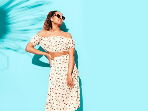 Hermosa mujer negra con peinado de rizos afro Modelo sonriente vestida con ropa hipster de verano Sexy mujer despreocupada posando junto a la pared azul en el estudio Bronceada y alegre En gafas de sol