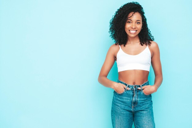 Hermosa mujer negra con peinado de rizos afro Modelo sonriente vestida con top blanco y jeans Mujer sexy despreocupada posando junto a la pared azul en el estudio Bronceada y alegre Aislado