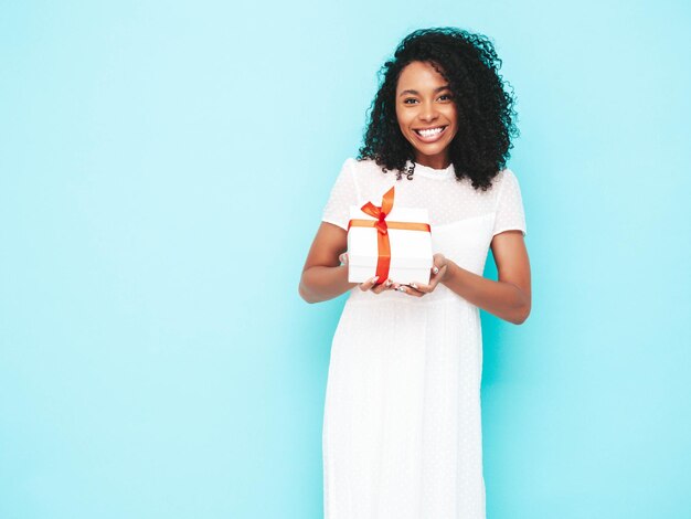 Hermosa mujer negra con peinado de rizos afro Modelo sonriente vestida con vestido blanco de verano Mujer sexy despreocupada posando cerca de la pared azul en el estudio Bronceada y alegre Caja de regalo de sujeción aislada