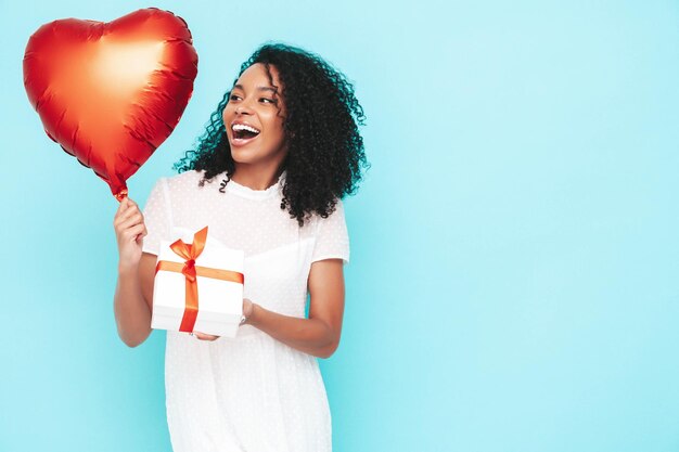 Hermosa mujer negra con peinado de rizos afro Modelo sonriente vestida con vestido blanco de verano Sexy mujer despreocupada posando cerca de la pared azul en el estudio Sosteniendo el globo de aire del corazón y la caja de regalo Aislado