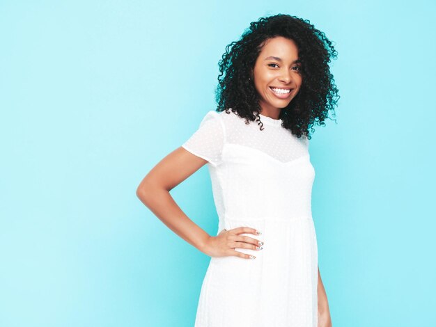 Hermosa mujer negra con peinado de rizos afro Modelo sonriente vestida con vestido blanco de verano Sexy mujer despreocupada posando junto a la pared azul en el estudio Bronceada y alegre Aislado