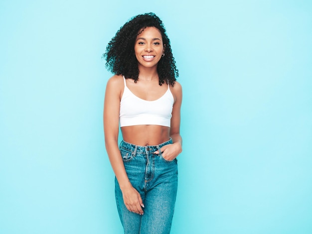 Hermosa mujer negra con peinado de rizos afro Modelo sonriente vestida con vestido blanco de verano Sexy mujer despreocupada posando junto a la pared azul en el estudio Bronceada y alegre Aislado