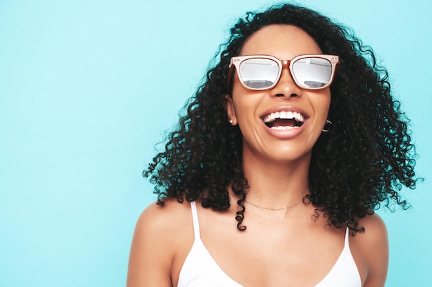 Hermosa mujer negra con peinado de rizos afro Modelo sonriente vestida con vestido blanco de verano Sexy mujer despreocupada posando junto a la pared azul en el estudio Bronceada y alegre En gafas de sol Primer plano