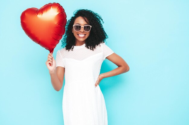 Hermosa mujer negra con peinado de rizos afro Modelo sonriente vestida con un vestido blanco de verano Sexy mujer despreocupada posando junto a la pared azul en el estudio Bronceada y alegre Sosteniendo un globo de aire del corazón