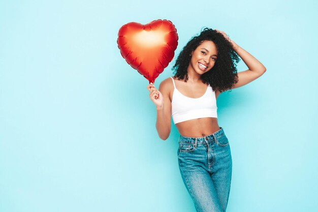 Hermosa mujer negra con peinado de rizos afro Modelo sonriente vestida con un vestido blanco de verano Sexy mujer despreocupada posando junto a la pared azul en el estudio Bronceada y alegre Sosteniendo un globo de aire del corazón