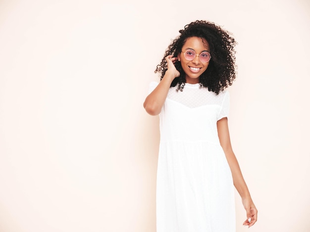 Hermosa mujer negra con peinado de rizos afro Modelo sonriente vestida con vestido blanco de verano Sexy mujer despreocupada posando junto a la pared en el estudio Bronceada y alegre Aislada