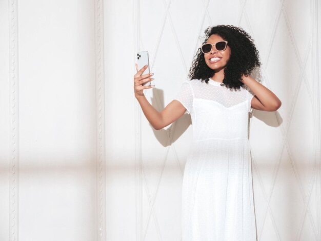 Hermosa mujer negra con peinado de rizos afro Modelo sonriente vestida con un vestido blanco de verano Sexy mujer despreocupada posando junto a la pared en el estudio En un día soleado Sombra desde la ventana Tomando fotos selfie