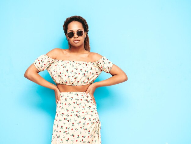 Hermosa mujer negra con peinado de rizos afro Modelo sonriente vestida con vestido de verano Sexy mujer despreocupada posando junto a la pared azul en el estudio Bronceada y alegre