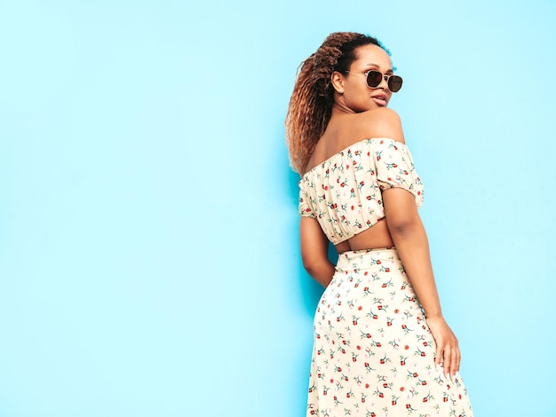 Hermosa mujer negra con peinado de rizos afro Modelo sonriente vestida con vestido de verano Sexy mujer despreocupada posando junto a la pared azul en el estudio Bronceada y alegre