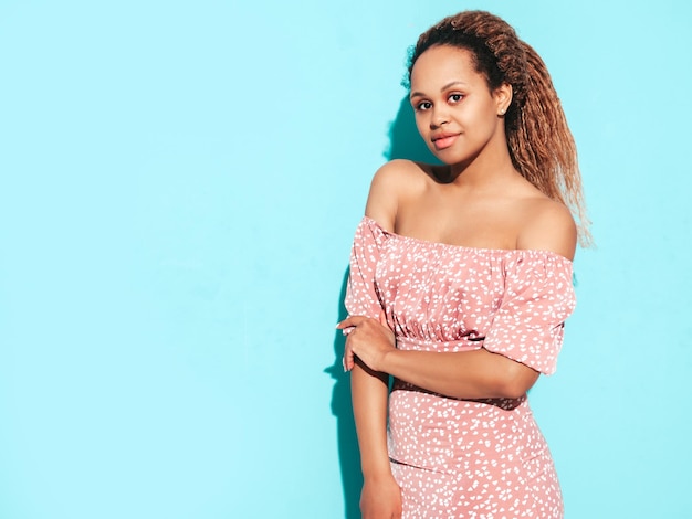 Foto gratuita hermosa mujer negra con peinado de rizos afro modelo sonriente vestida con vestido de verano sexy mujer despreocupada posando junto a la pared azul en el estudio bronceada y alegre