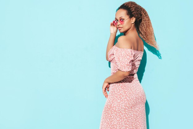 Hermosa mujer negra con peinado de rizos afro Modelo sonriente vestida con vestido de verano Sexy mujer despreocupada posando junto a la pared azul en el estudio Bronceada y alegre