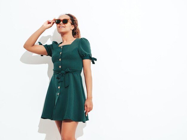 Hermosa mujer negra con peinado de rizos afro Modelo sonriente vestida con un vestido verde de verano blanco Mujer despreocupada sexy posando junto a la pared en el estudio Bronceada y alegre Aislada en gafas de sol