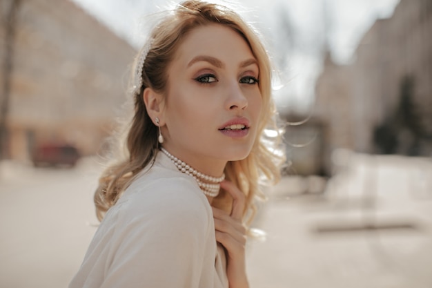 Hermosa mujer de ojos grises con maquillaje mira a la cámara. Chica guapa en blusa blanca y collar de perlas posa fuera