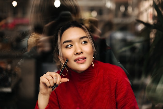 Hermosa mujer de ojos marrones en grandes aretes de oro y traje rojo posando pensativamente con lápiz en sus manos