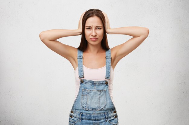 Hermosa mujer en overoles de mezclilla que sufren de fuertes ruidos y dolor de cabeza, cubriendo la cabeza y las orejas con las manos, mirando molesto aislado. Emociones humanas, sentimientos y reacciones