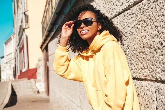 Hermosa mujer con peinado de rizos afro Modelo hipster sonriente con capucha amarilla Mujer despreocupada sexy posando en el fondo de la calle con gafas de sol Alegre y feliz