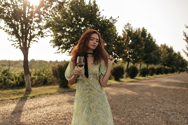 Hermosa mujer con peinado rojo esponjoso y venda negra en el cuello con elegante vestido de verano sosteniendo un vaso con vino y sombrero al aire libre