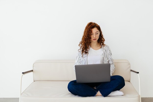 Hermosa mujer pelirroja que trabaja en línea usando una computadora portátil sentada en un cómodo sofá gris