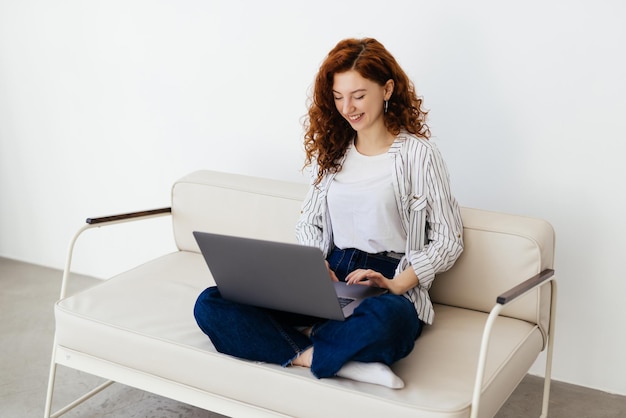 Hermosa mujer pelirroja que trabaja en línea usando una computadora portátil sentada en un cómodo sofá gris