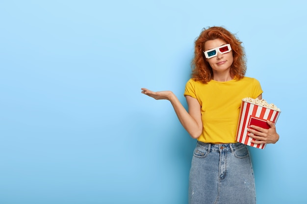 Hermosa mujer pelirroja usa lentes virtuales, camiseta amarilla y falda de mezclilla, sostiene una canasta de palomitas de maíz, entra al cine, tiene expresión dudosa, duda qué película elegir para ver.