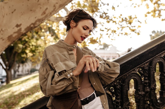 Hermosa mujer con pelo corto en chaqueta verde oliva mira el reloj al aire libre. Mujer morena con bolso con labios brillantes posa afuera.