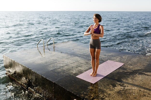 Hermosa mujer con pelo corto oscuro de pie con los ojos cerrados y meditando junto al mar. Bella dama en top deportivo y pantalones cortos haciendo yoga con vista al mar en el fondo