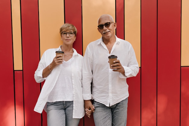 Hermosa mujer con el pelo corto en ropa ligera posando con una taza de café y sosteniendo la mano con el hombre de pelo gris en rojo y naranja.