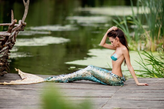 Hermosa mujer con pelo largo y vestida como una sirena se sienta en el puente sobre el agua
