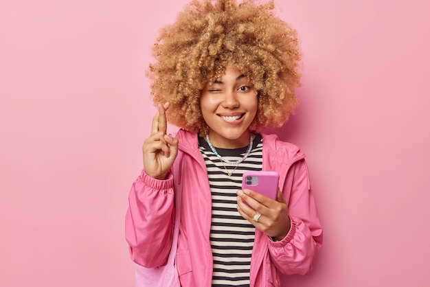 Foto gratuita hermosa mujer de pelo rizado esperanzada sostiene el teléfono móvil mantiene los dedos cruzados cree en la buena suerte guiña los ojos vestida con una chaqueta informal aislada sobre fondo rosa que mis sueños se hagan realidad