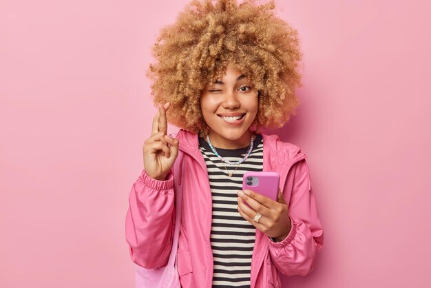 Hermosa mujer de pelo rizado esperanzada sostiene el teléfono móvil mantiene los dedos cruzados cree en la buena suerte guiña los ojos vestida con una chaqueta informal aislada sobre fondo rosa Que mis sueños se hagan realidad