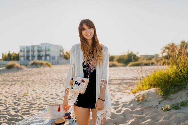 Hermosa mujer con pelos morenos ondulados, vestida de blanco boho cover up mira a la cámara con una sonrisa. Posando en la playa cerca del hotel. Colores del atardecer.