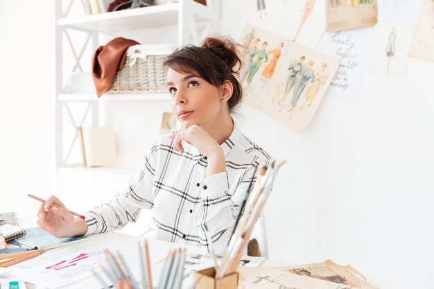 Hermosa mujer pensativa diseñadora de modas sentada en su lugar de trabajo