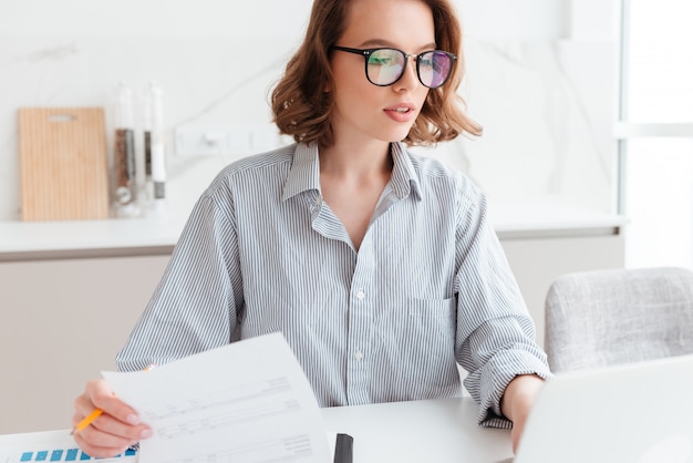 Hermosa mujer pensativa en gafas y camisa a rayas trabajando con documentos en casa