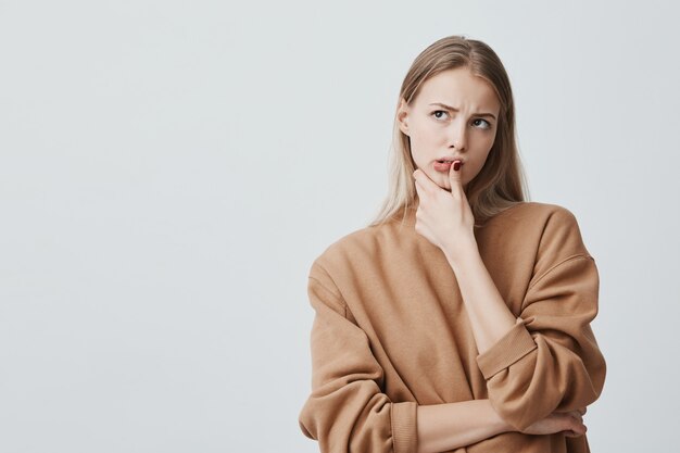 hermosa mujer pensativa con largo cabello rubio, mira hacia arriba con expresiones pensativas, planea algo, posa contra la pared en blanco. Grave hembra concentrada manteniendo el dedo en la barbilla