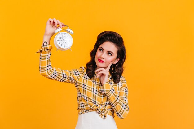 Hermosa mujer pensativa con reloj. Foto de estudio de juguetona chica pinup posando sobre fondo amarillo.