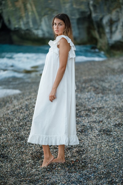 Foto gratuita hermosa mujer de pie y mirando a la orilla del mar en vestido blanco durante la puesta de sol y mirando triste. .