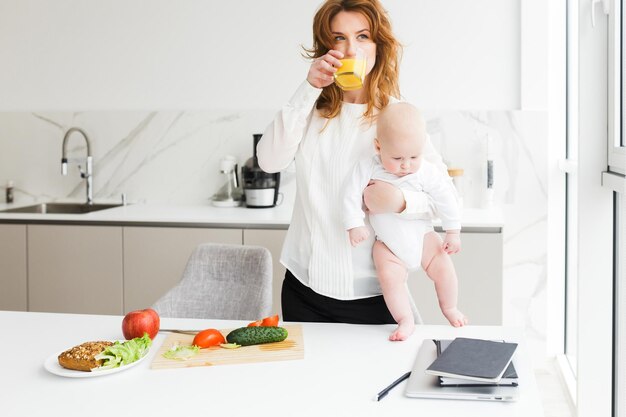 Hermosa mujer de pie y sosteniendo a su lindo bebé mientras bebe jugo y cocina en la cocina