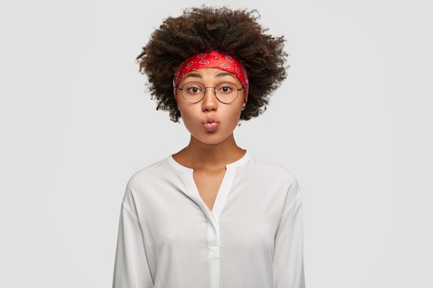 Hermosa mujer de piel oscura hace pucheros en los labios, tiene corte de pelo afro, hace muecas, usa anteojos redondos, diadema roja y camisa blanca, está de pie contra la pared. concepto de expresiones faciales