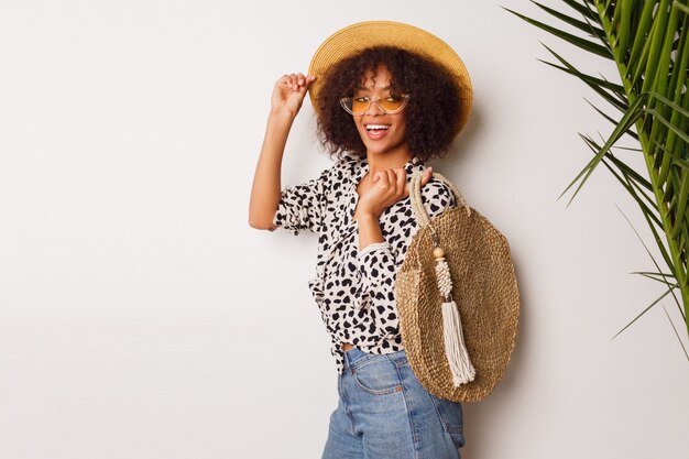 Hermosa mujer con piel oscura en jeans y sombrero de paja posando en estudio sobre fondo blanco con bolsa en estilo Bali. Sipping humor.