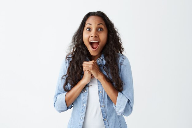 Hermosa mujer de piel oscura en ropa casual con la boca abierta celebrando su victoria manteniendo juntos los puños cerrados con expresión de emoción y satisfacción. Mujer regocijándose de su éxito.