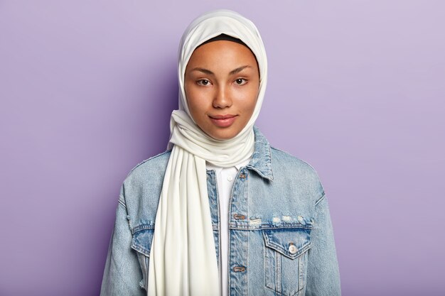 Hermosa mujer con piel oscura sana, viste hijab blanco, vestida con chaqueta de mezclilla, mira directamente, aislada sobre pared púrpura. La mujer religiosa tiene una belleza natural. Concepto de etnia