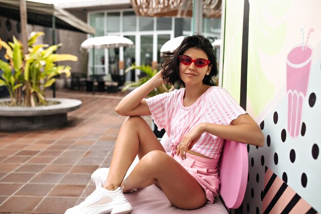 Hermosa mujer posando en la cafetería al aire libre. Mujer joven con estilo en gafas de sol rosas mirando a cámara.